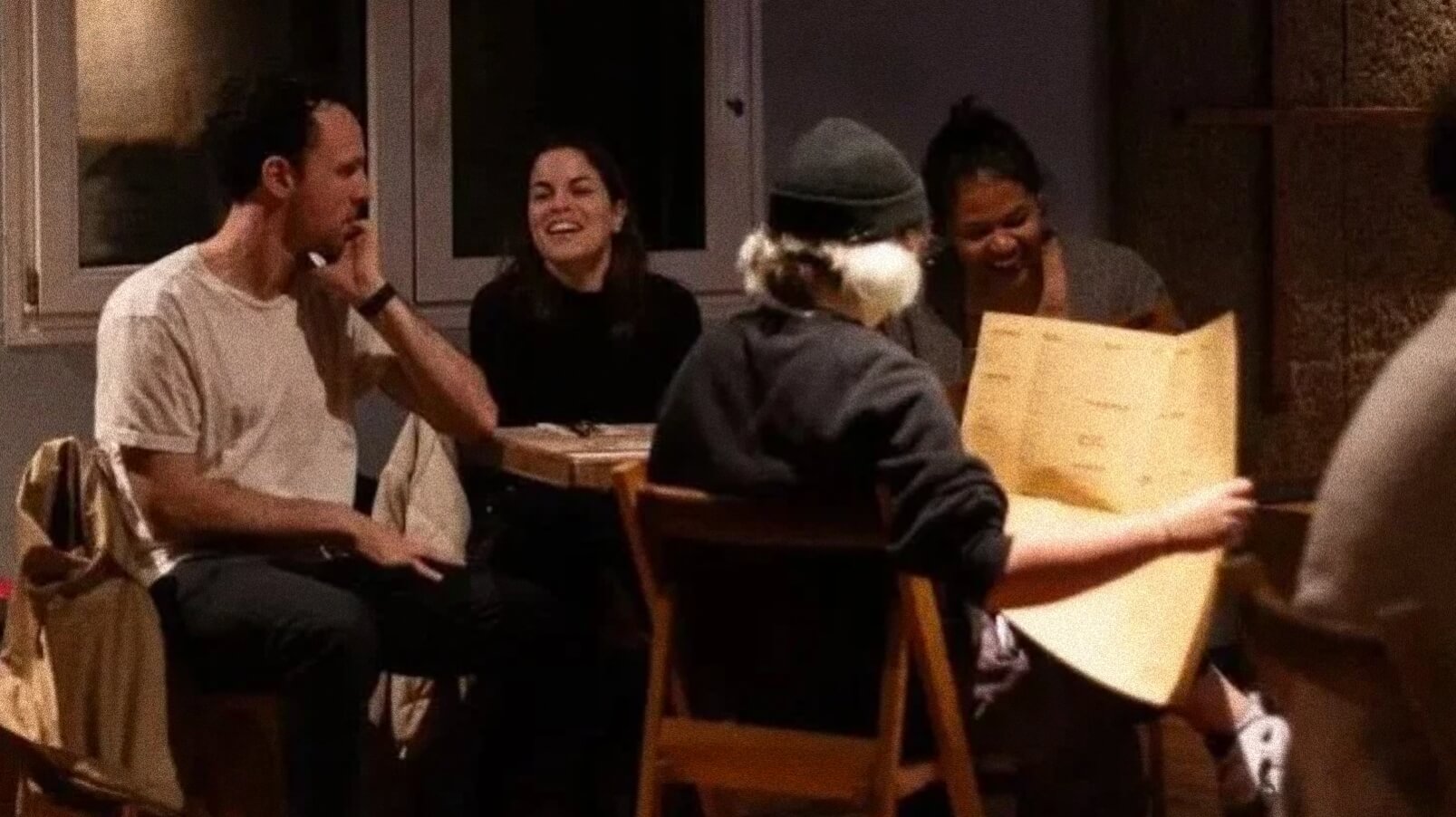 Four people sitting at a restaurant table, engaging in conversation, with two women smiling and two men, one wearing a beanie, looking at a menu.