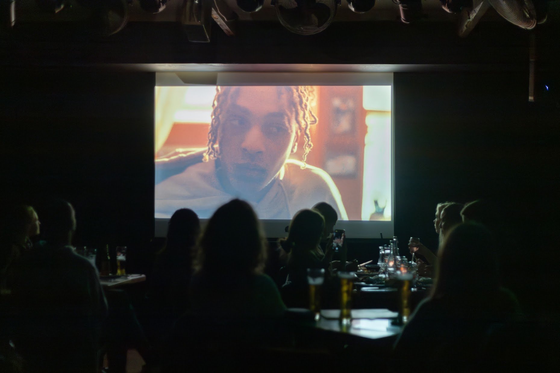 People sitting in a dimly lit room watching a large screen showing a close-up of a person's face, with drinks on the tables.