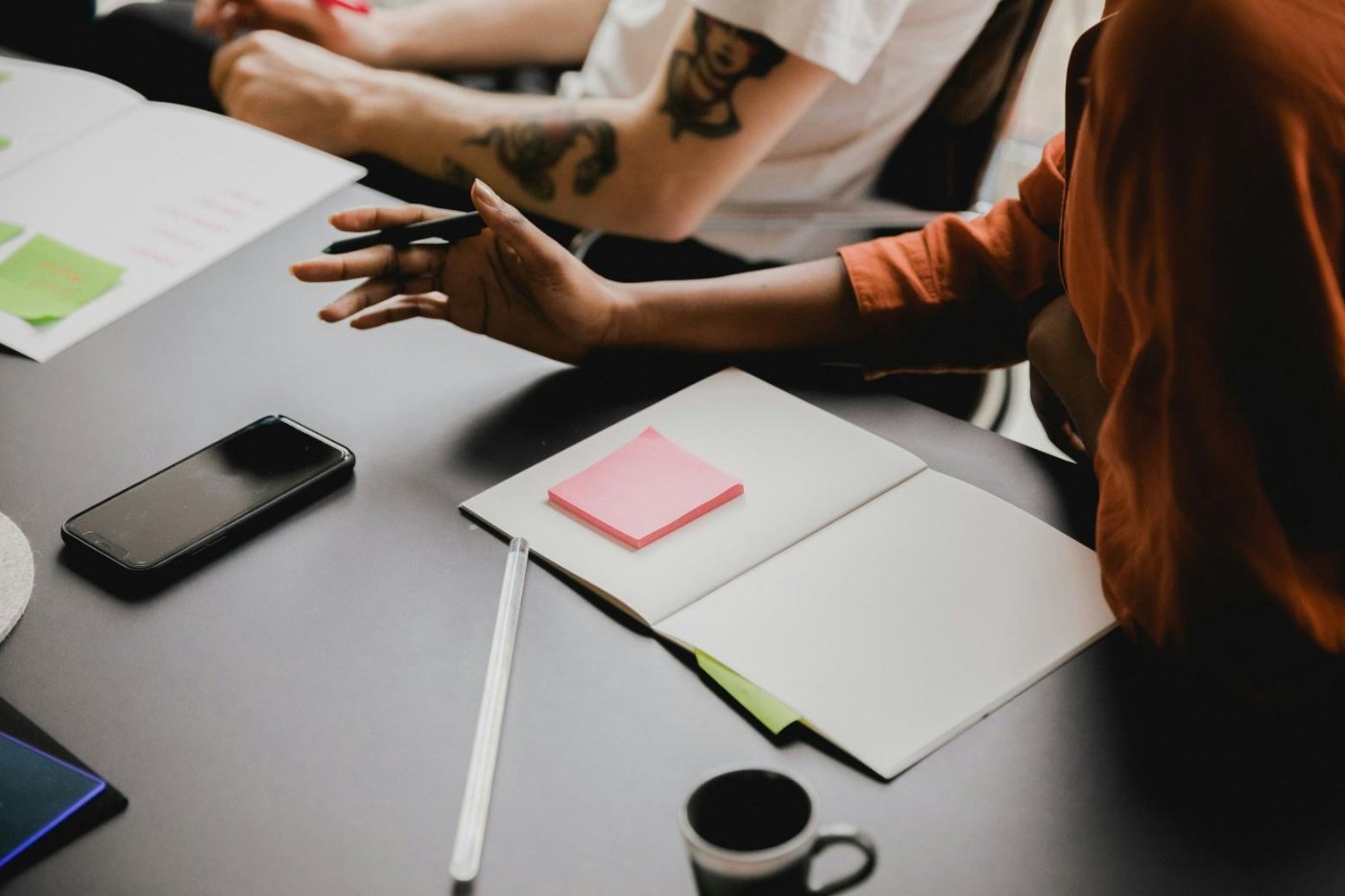 People sitting around a table with notebooks, sticky notes, a smartphone, a pen, and a coffee mug.