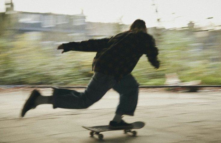 Person skateboarding outdoors on a blurry background with trees and buildings.