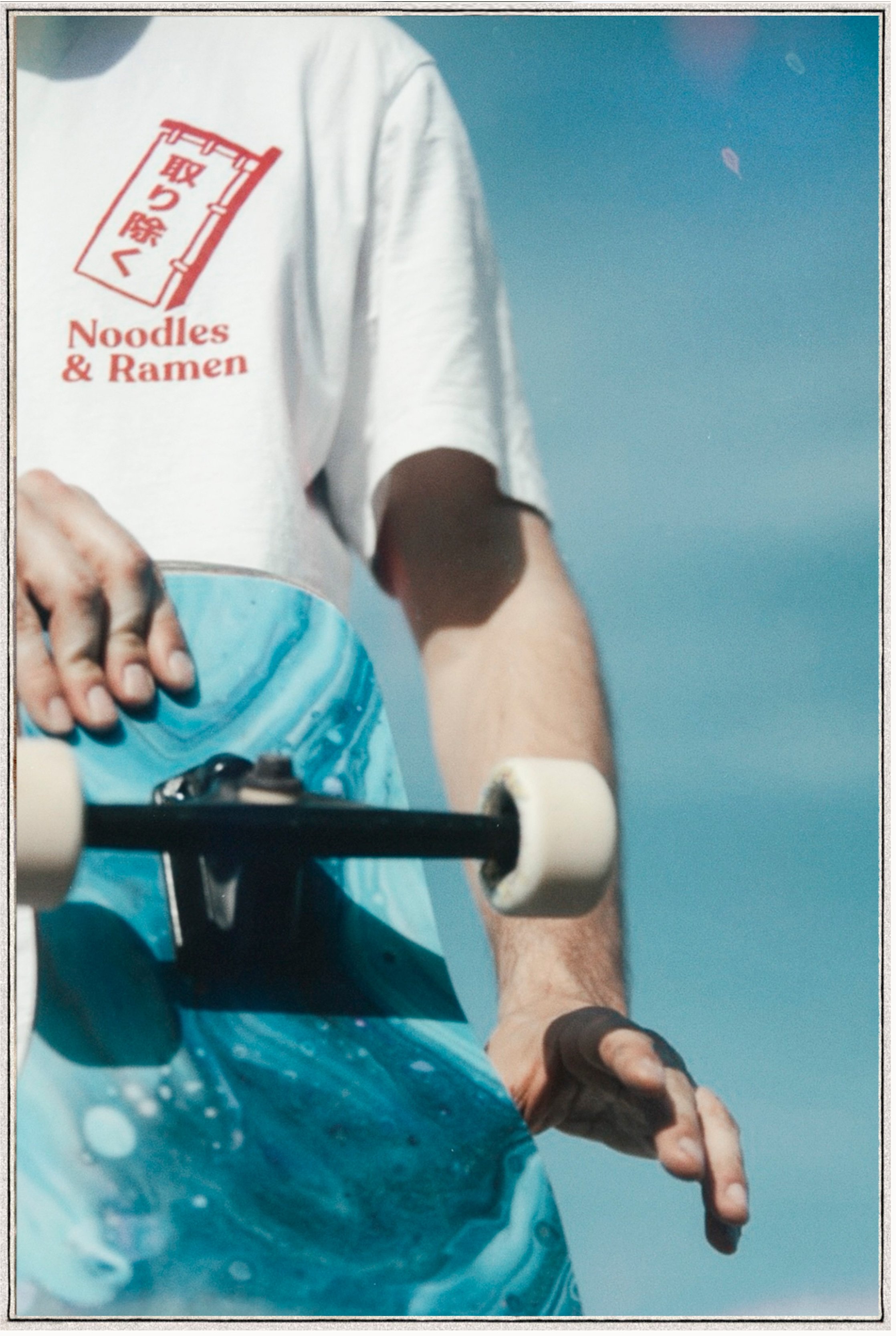 Close-up of a skateboarder wearing a white T-shirt with Japanese characters and English text, holding a skateboard, against a blue sky background.