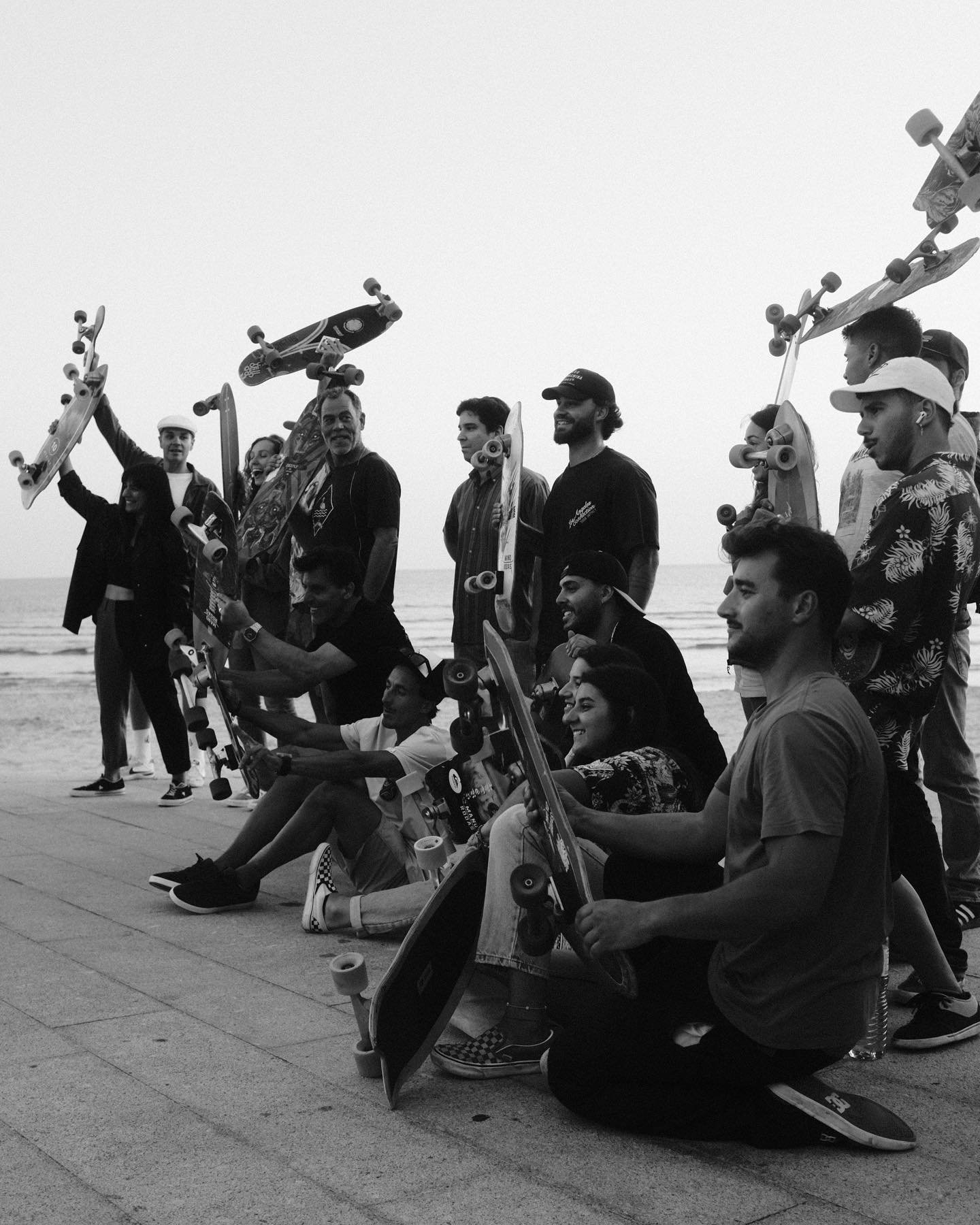 A group of people at the beach holding skateboards, some standing and some sitting, celebrating together with a foggy sky and ocean in the background.