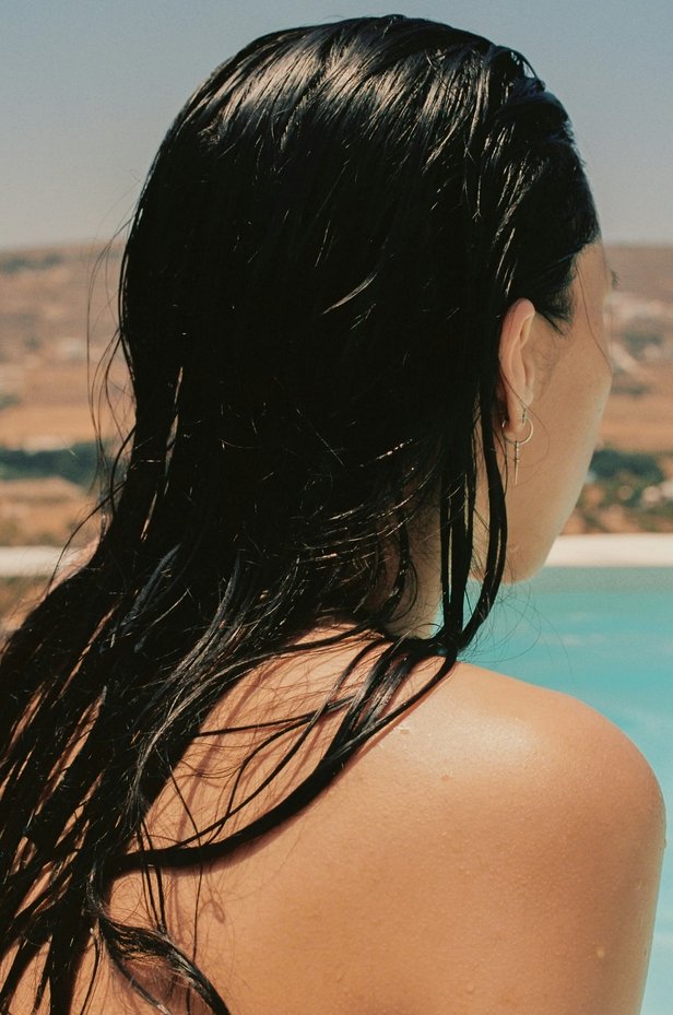 A woman with wet black hair, wearing a hoop earring, looking away from the camera near a pool, with a desert landscape in the background.