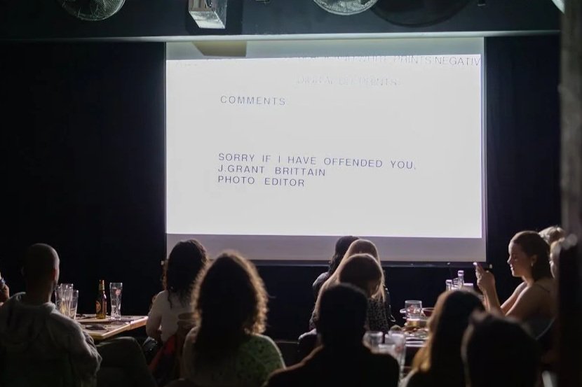 Audience watching a presentation screen displaying a message that says, 'Sorry if I have offended you. J. Grant Brittain. Photo Editor.'