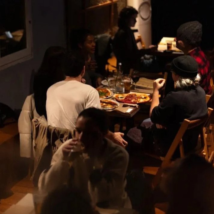 A group of people dining together in a dimly-lit restaurant, with some eating pizza and others engaging in conversation.