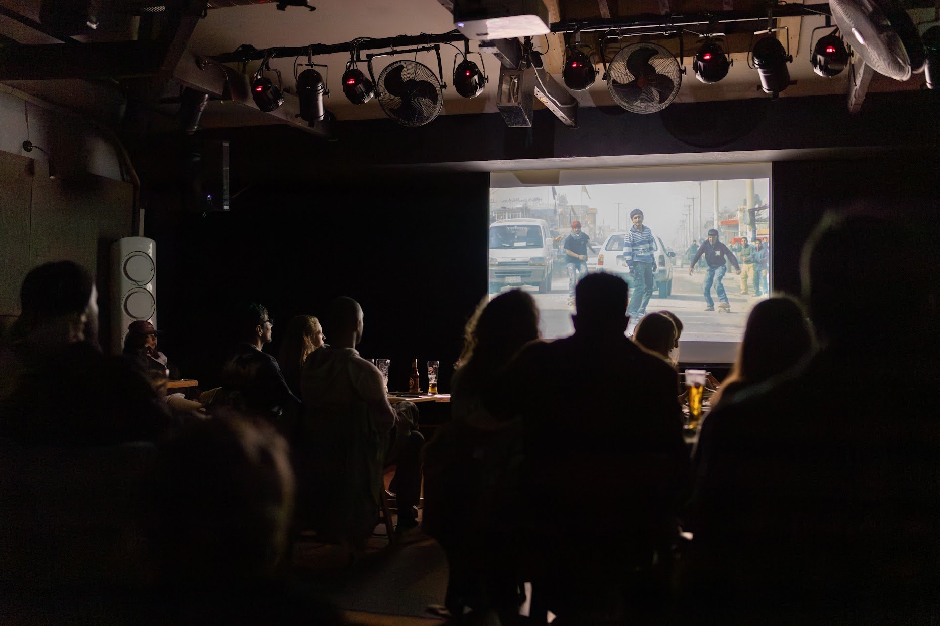 People watching a movie or video on a large screen in a dark room, with some seated at tables with drinks, possibly in a bar or theater setting, with lights and fans mounted on the ceiling.