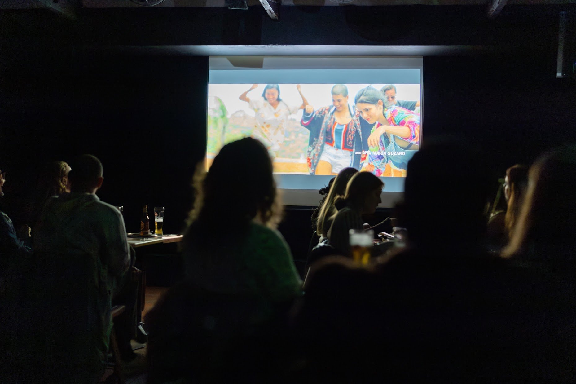 People sitting in a dark room watching a movie or presentation on a large screen, with some holding drinks.