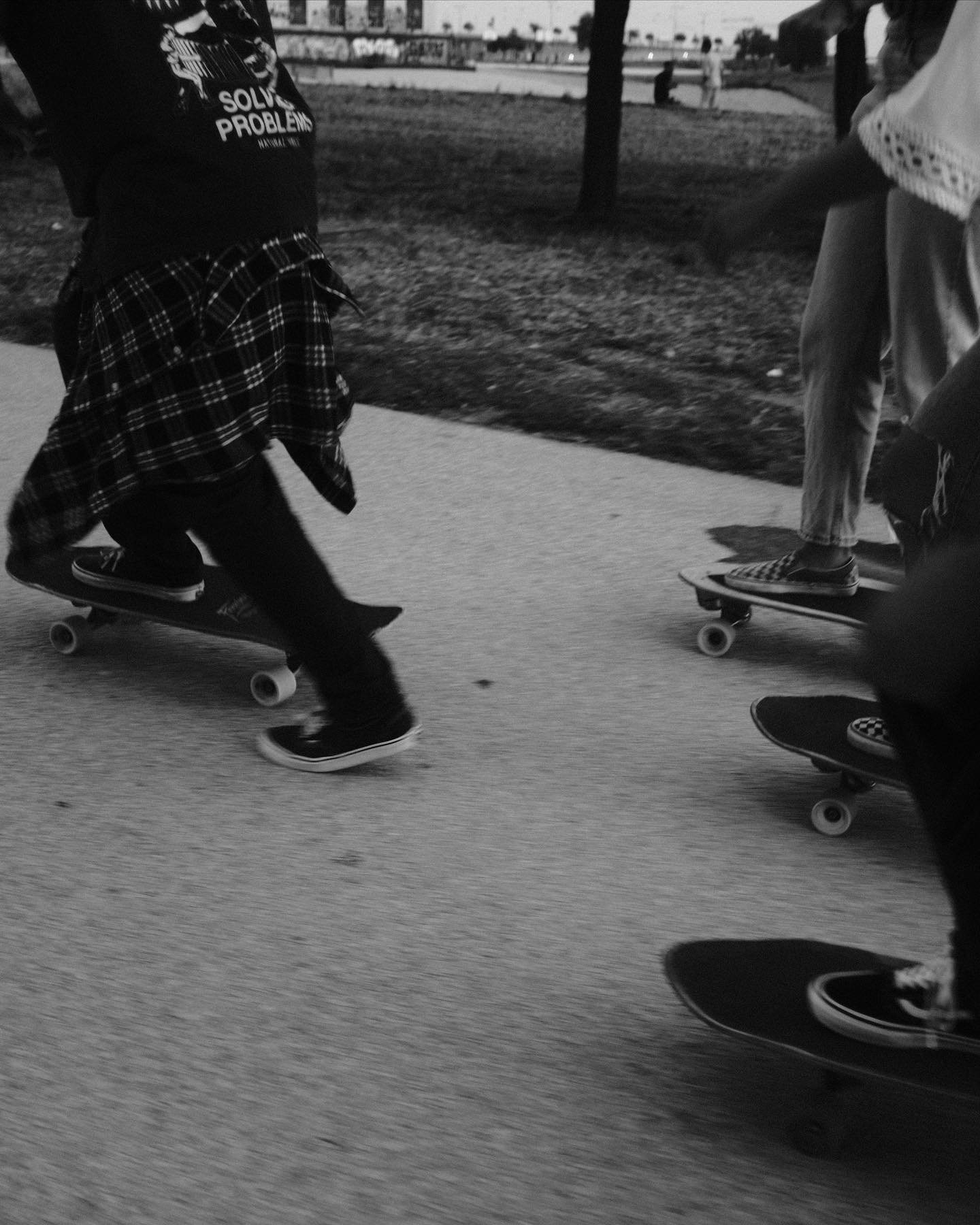People skateboarding on a paved path in a park, with some trees and a playground in the background.