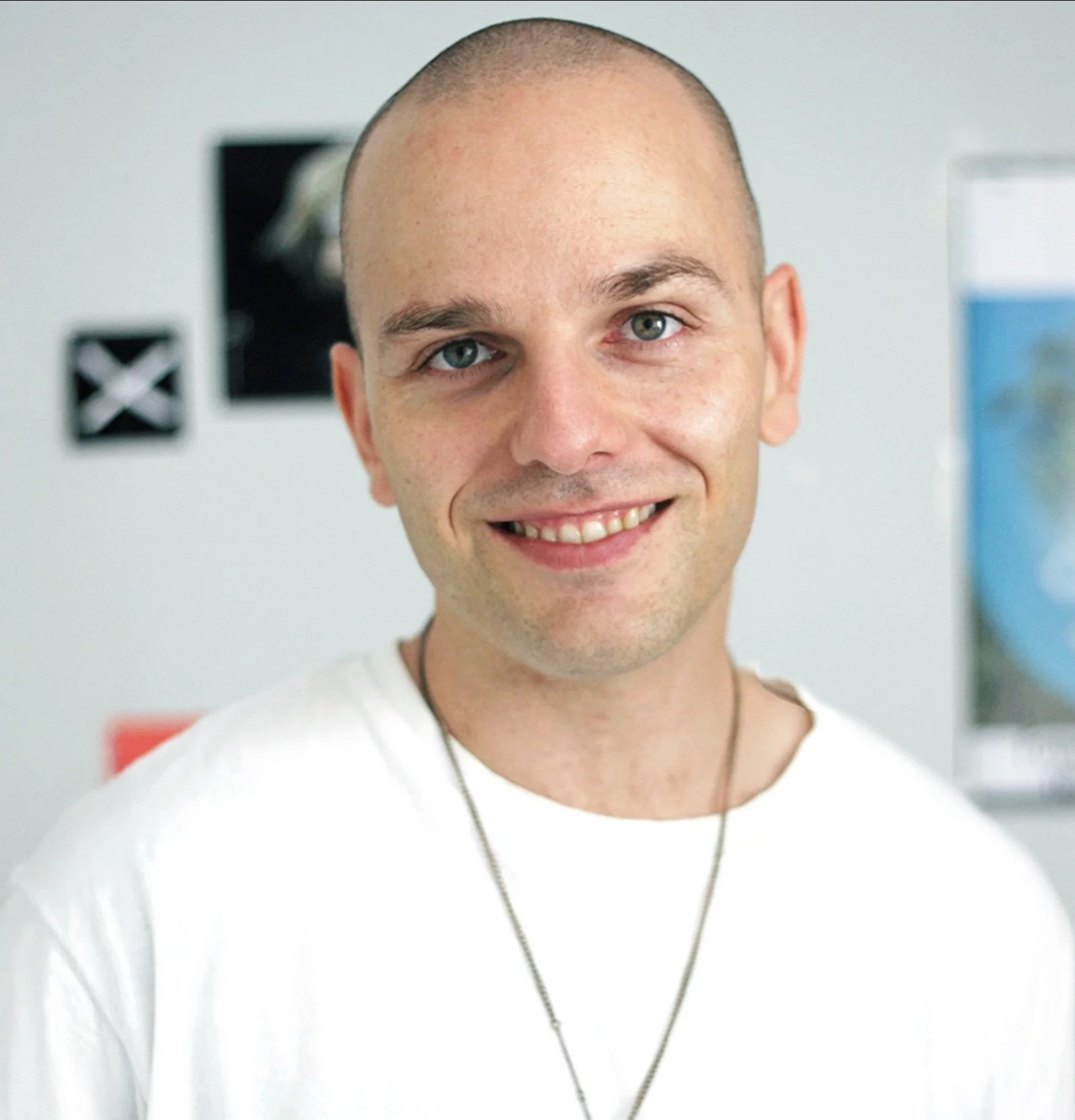Smiling young man with a shaved head and blue eyes wearing a white shirt and necklace, standing indoors with posters on the wall behind him.