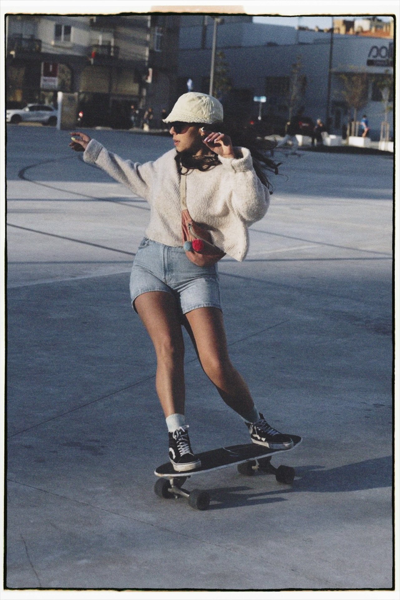 A woman skateboarding at a skate park, wearing a white beanie, sunglasses, a beige sweater, denim shorts, and black sneakers.