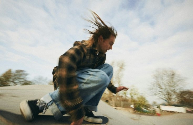 Person crouching on skateboard on a slope outdoors, hair blowing in the wind, with trees and sky in background.