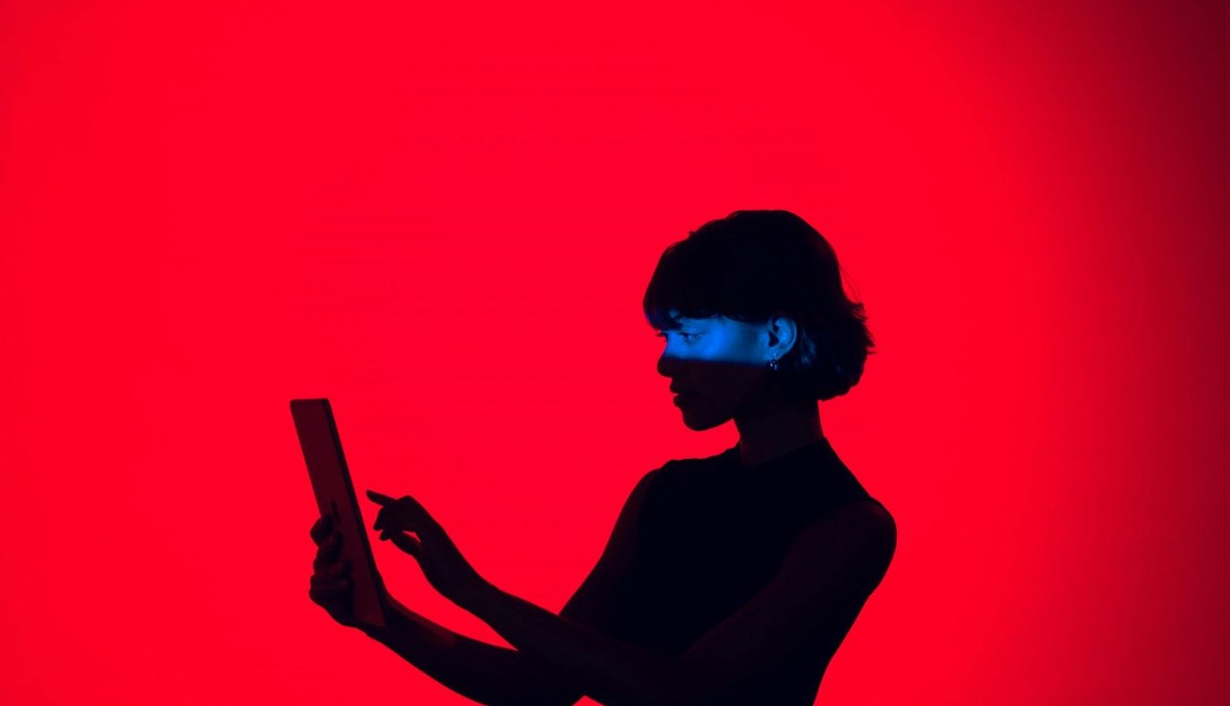 Silhouette of a woman with short hair using a tablet against a red background, with a blue light on her face.