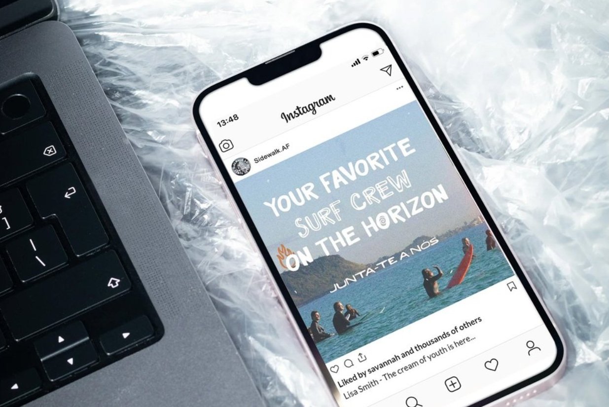 Smartphone displaying an Instagram post about surf crew on the horizon, showing people surfing in the ocean with a mountain in the background, placed beside a laptop keyboard on a plastic surface.