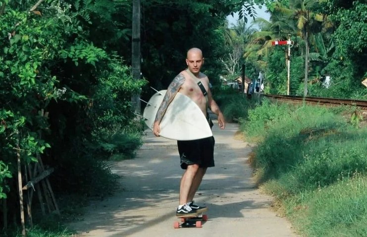 A man with tattoos and no shirt is skateboarding on a dirt path with a surfboard under his arm, surrounded by green trees and bushes, near a railroad track.