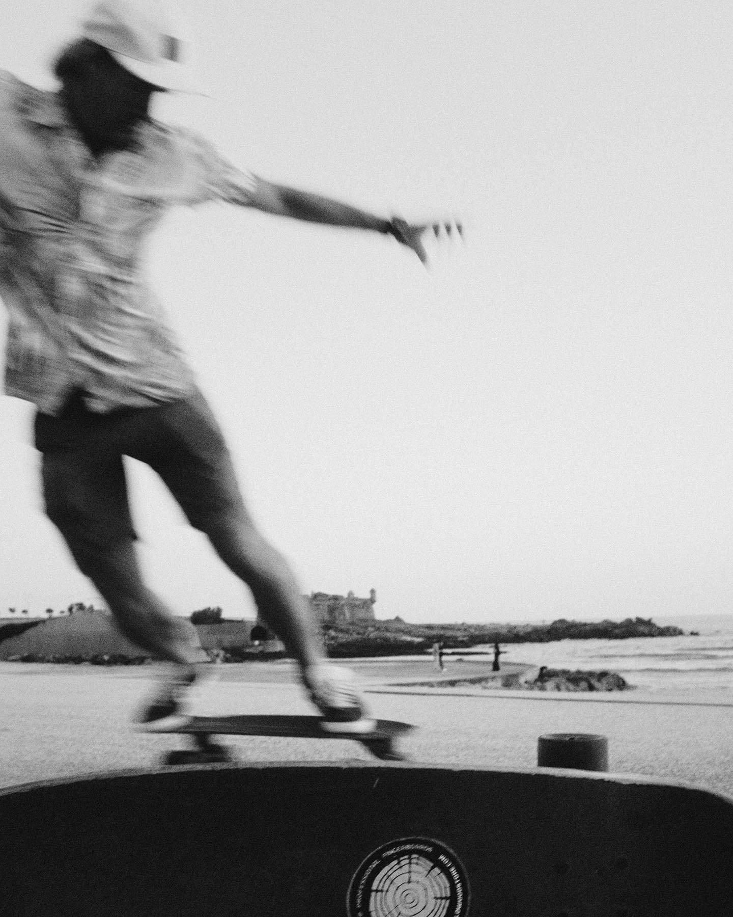 A person skateboarding on a beach at sunset, with the ocean and a small island in the background, while wearing a cap and casual clothes.