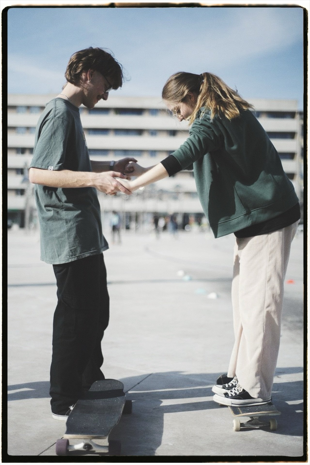 Two young people, a male and a female, are at a skate park. The male is standing on a skateboard and wearing a dark t-shirt and glasses. The female is on a skateboard, wearing a green jacket and beige pants. She is helping the male, possibly adjustin