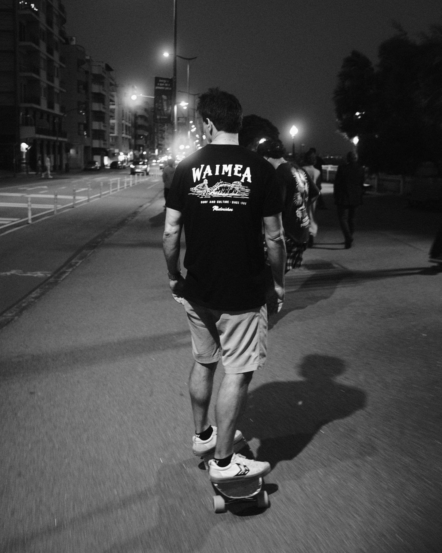 Young man skateboarding on a city street at night, wearing a T-shirt with "WAIMEA" written on the back.
