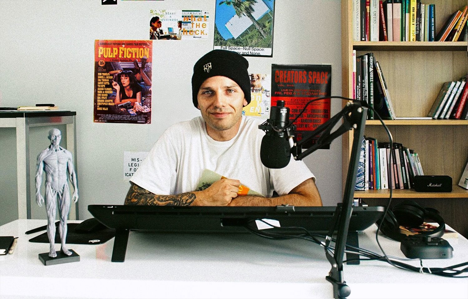 A man sitting at a desk in a room with posters on the wall and a bookshelf behind him, with a microphone in front of him.