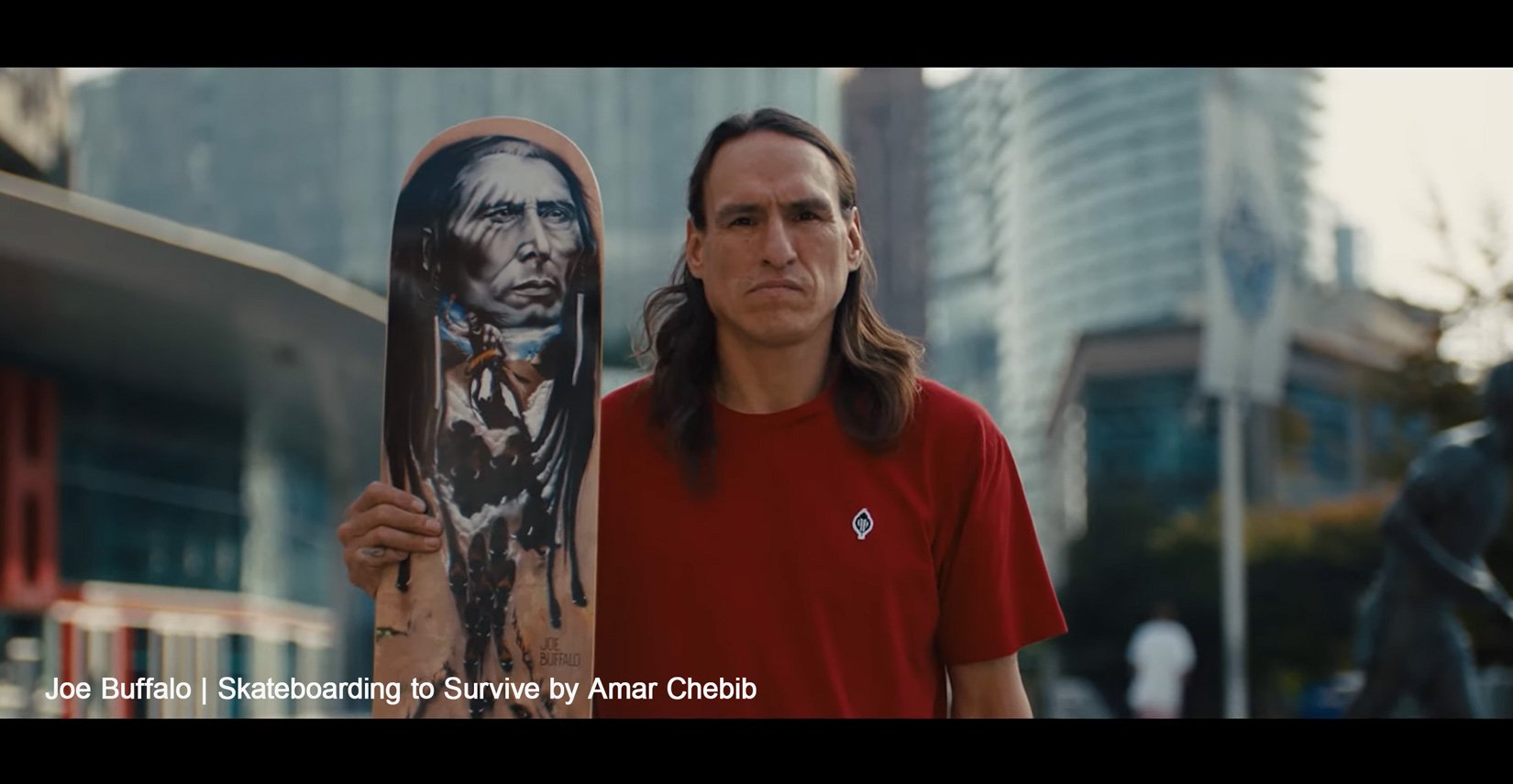 A man with long hair wearing a red t-shirt holding a skateboard with a Native American portrait illustration on it, in an urban area with modern buildings in the background.
