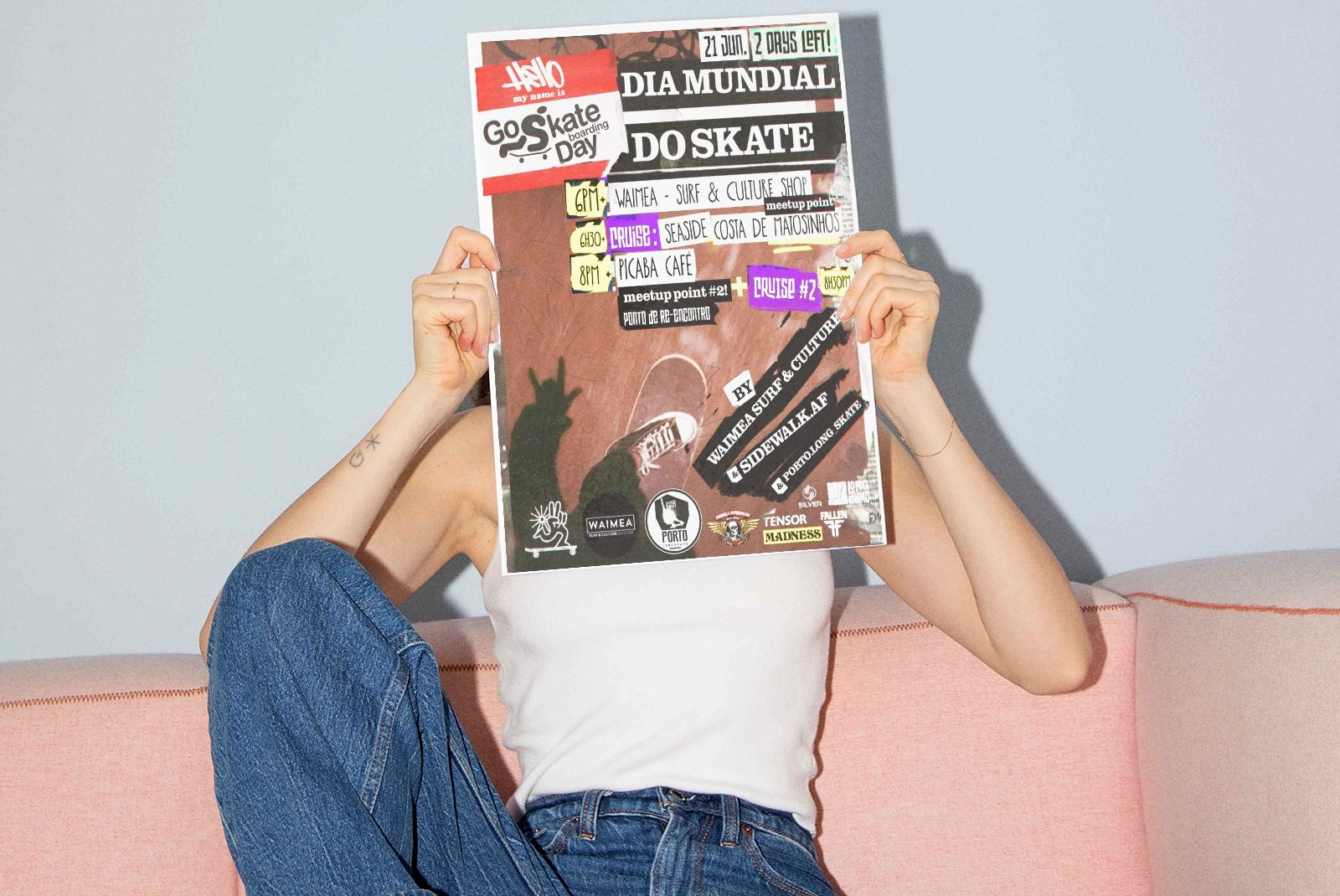 Person sitting on a pink couch holding a colorful event flyer in front of their face, wearing a white tank top and blue jeans, with tattoos on their arm, and a bracelet on their wrist.