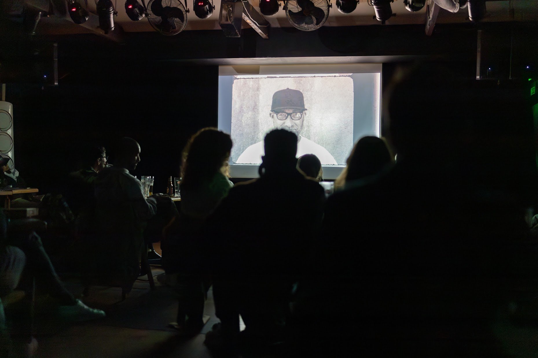 People watching a projected video of a man wearing glasses and a baseball cap in a dark room, possibly a nightclub or lounge.