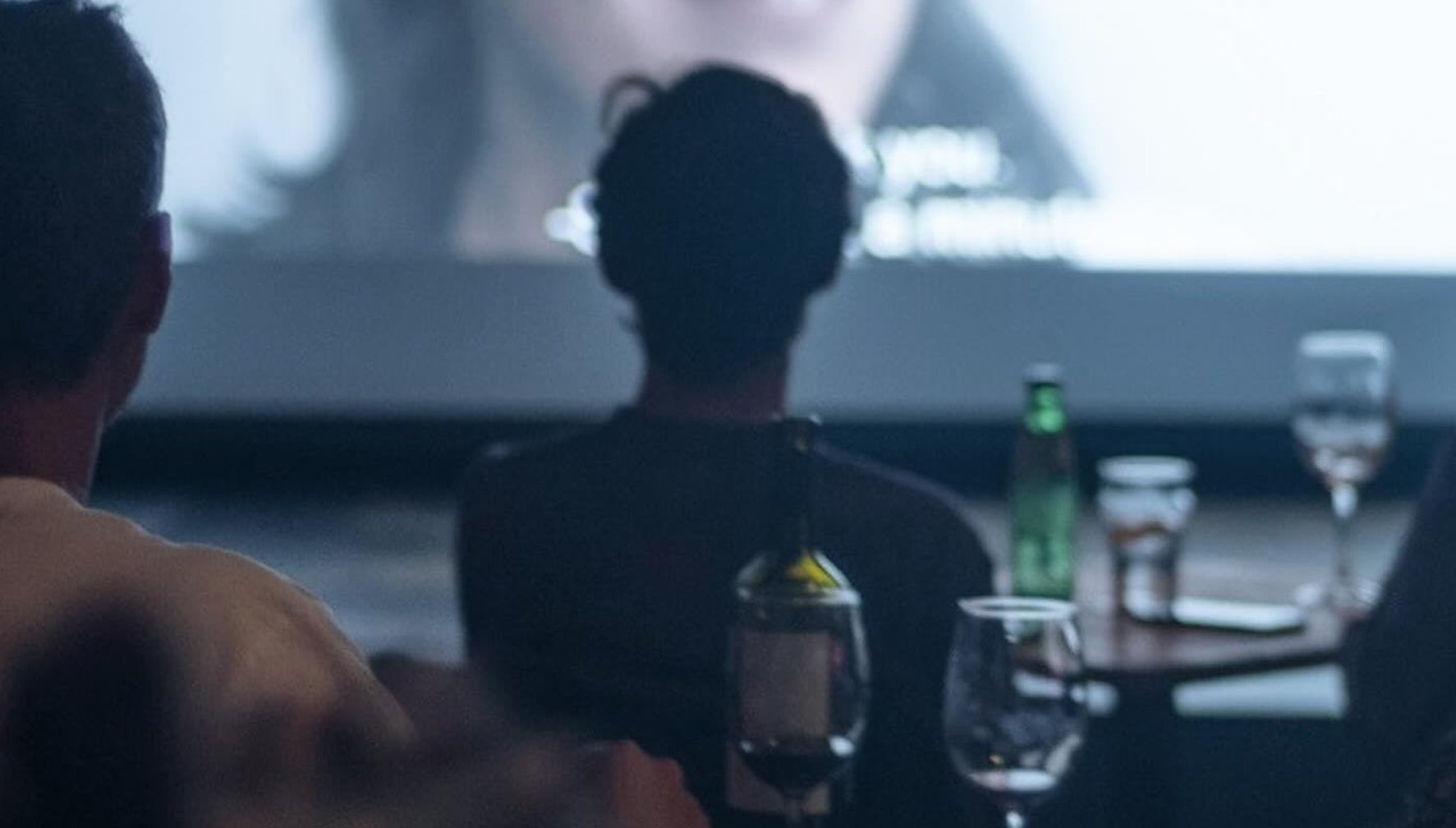 People watching a movie in a dark theater, with drinks and bottles on a table in front of them.