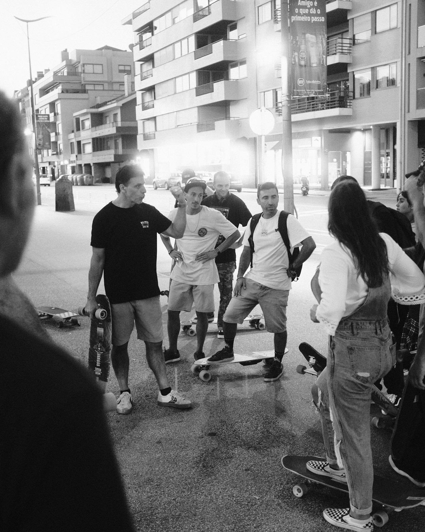 A group of young people with skateboards gathered on a city street, listening to a man in a black shirt speaking.