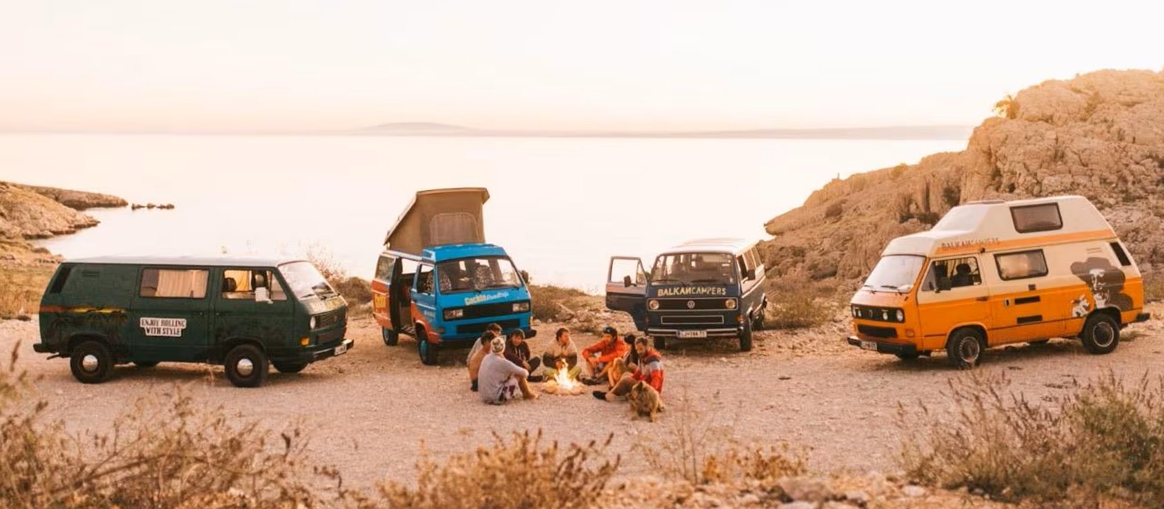 A group of people sitting around a campfire near four colorful camper vans by the water's edge at sunset, surrounded by rocky terrain and sparse vegetation.