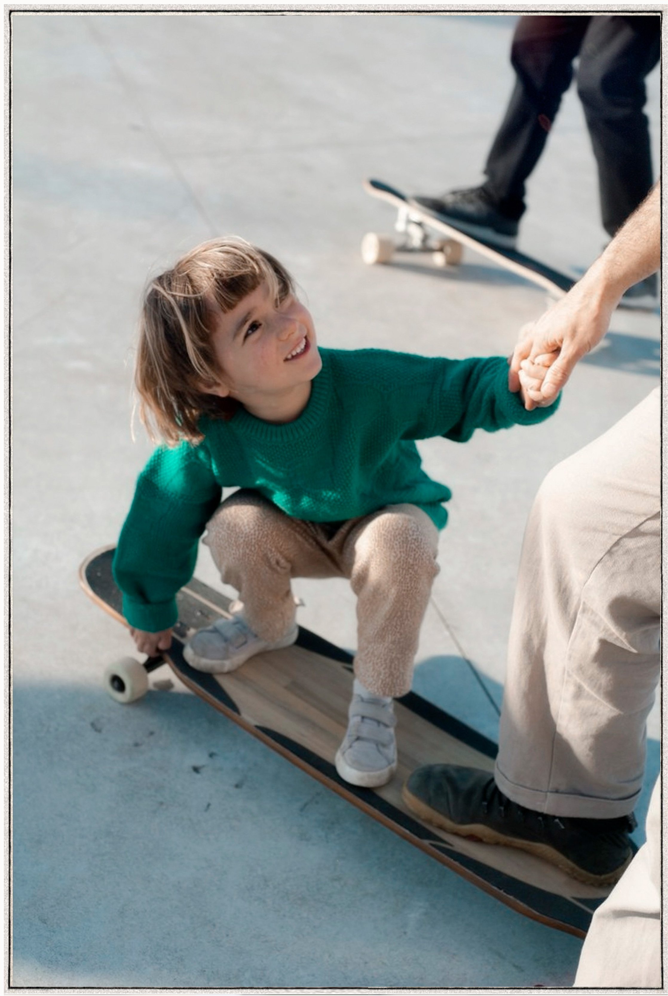A young child with a green sweater riding a skateboard, smiling, with an adult holding their hand for support.