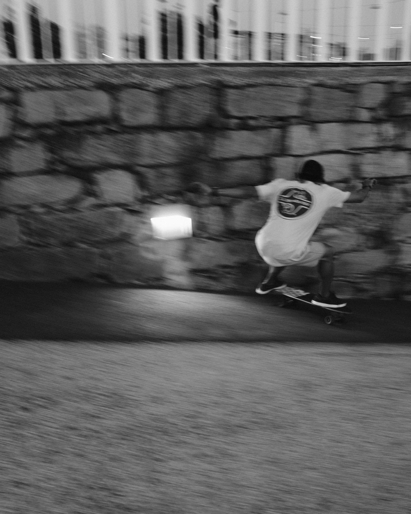 A person skateboarding along a street at night, wearing a t-shirt with a circular logo on the back, next to a brick wall.