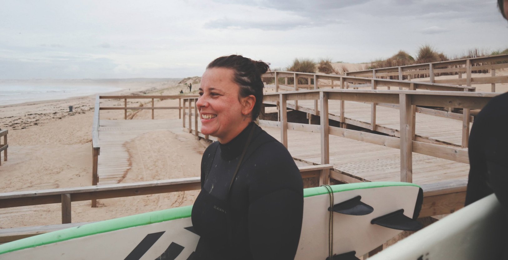 A smiling woman holding a surfboard on a beach with wooden walkways and dunes.