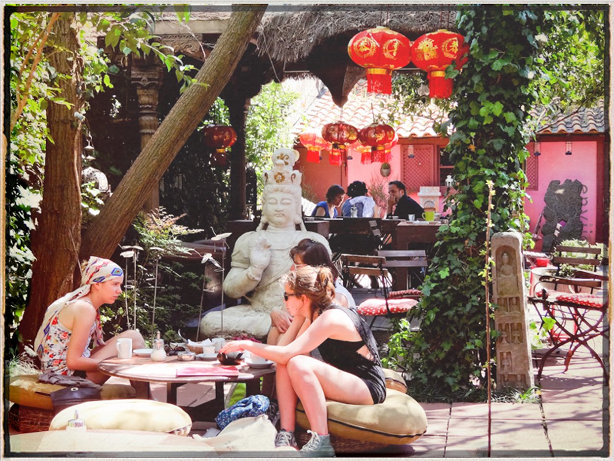 Outdoor café with Asian decorative elements, including red lanterns, a white stone statue, and a pink wall. People are sitting at tables, enjoying drinks and conversation.