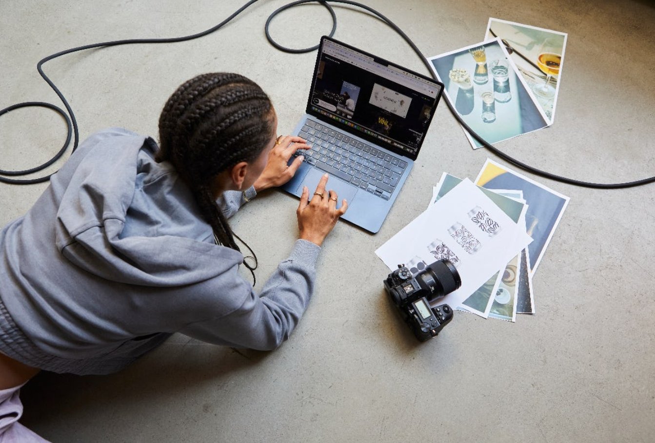 A woman with braided hair lies on the floor using a laptop, surrounded by printed photos, a camera, and external cables.