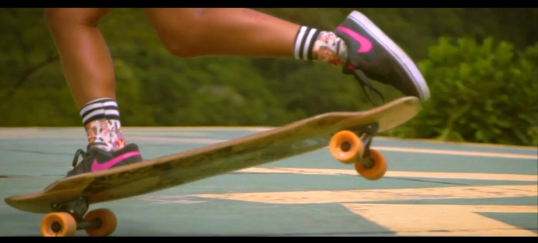 Close-up of a skateboarder’s legs and feet on a skateboard, wearing floral socks and black sneakers with pink accents, skating on a painted outdoor court.